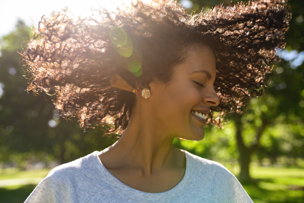 Woman smiling outdoors with sunlight highlighting her curly hair.